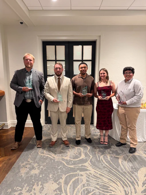 Five men and women standing indoors holding glass awards, dressed in formal and semi-formal attire, posing for a group photo in front of black double doors.