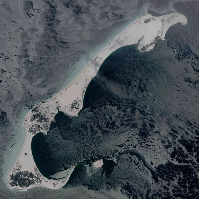 Satellite view of a long, curved sandy island surrounded by dark water and ice patterns.