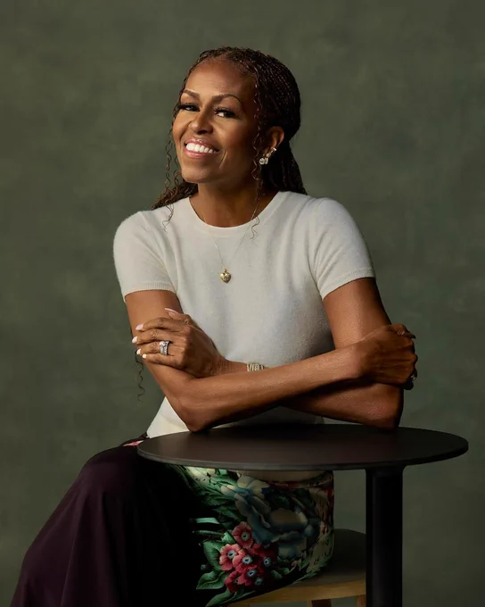 A photo of Michelle Obama set against a dark green background, seated at a small table and smiling.