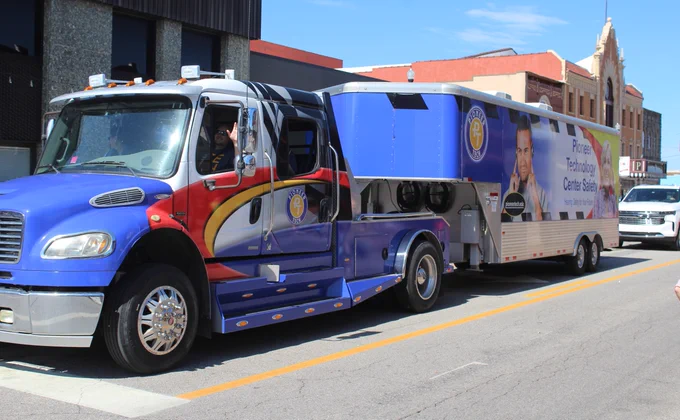 Blue and red semi-truck parked on a city street, towing a large trailer with a promotional image of a man and text on its side.