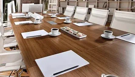A meeting room with a large wooden table and cream coloured charis.  The table is set with writing pens and pads and coffee and cake refreshments.