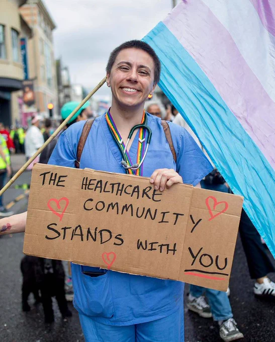 A healthcare worker holds a transgender flag at a rally while holding a sign that says "The healthcare community stands with you."