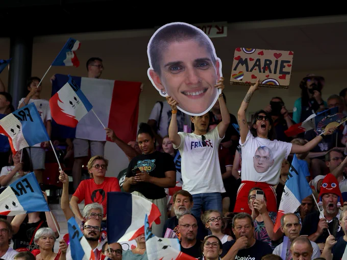 Marie Patouillet's loved ones at the cycling velodrome in the stands surrounded by French flags. They are holding a giant sign of her face and a cardboard sign that says Marie with red hearts, rainbows, and rainbow hearts