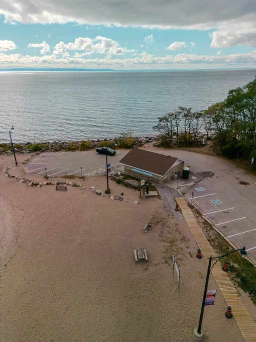 A sandy beach with a small building near the shoreline and calm water under a partly cloudy sky. A building housing public washrooms surrounding by parking spaces is in clear view.