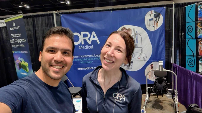 Varun Chandak (left) poses with Sarah Lambert, CEO of Ora Medical, at Abilities Expo Chicago. They stand in front of the Ora Medical exhibition booth, which features displays about pediatric mobility solutions and adaptive equipment. A mobility device is visible beside the booth, while additional accessibility and assistive technology exhibitors can be seen in the surrounding expo hall. 