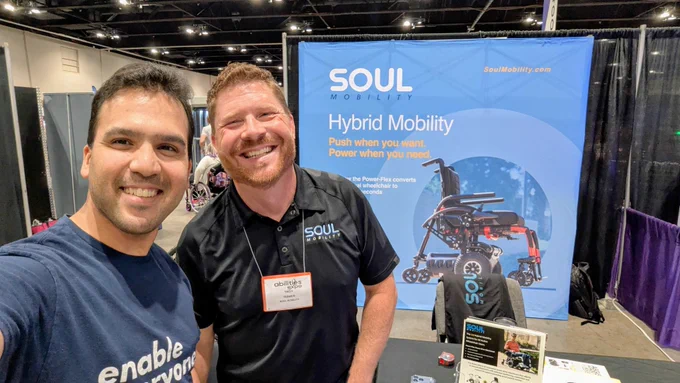 Varun Chandak (left) poses with Troy Tesmer, CEO of Soul Mobility and an ATS alumnus, at Abilities Expo Chicago. They stand in front of the Soul Mobility booth, which features information about hybrid mobility technology and a powered wheelchair solution. Product displays, promotional materials, and accessibility technology exhibits are visible throughout the expo hall.