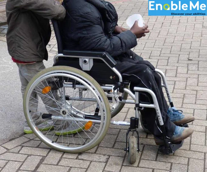 A person wearing a dark jacket sits in a manual wheelchair on a paved sidewalk while another person stands behind, appearing to assist by pushing the chair. The wheelchair has large rear wheels, small front casters, and footrests supporting the seated person's feet. The seated person is holding a white object in their hands. An "EnableMe" logo with the tagline "Discover your abilities" appears in the upper-right corner.