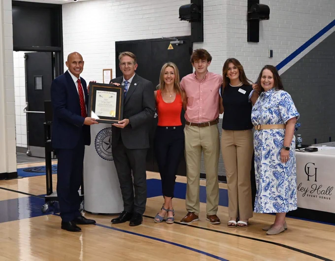Six adults pose in a gym, with two holding a framed “State of North Carolina” certificate while four others stand beside them.
