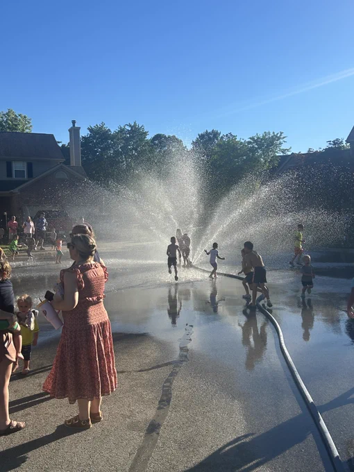 Children run and play through streams of water spraying from a fire hose during a Fishers Fire Department Cul-de-sac Drill. Families gather along the street as water creates a large mist under a clear blue sky.