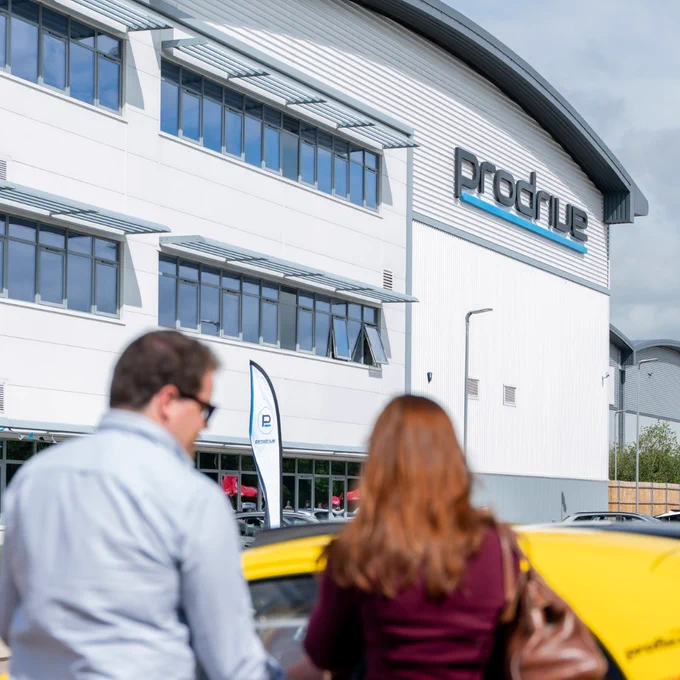 Exterior of Prodrive HQ in Banbury on a bright sunny day, with the Prodrive logo on the building. Two attendees walk past a yellow sports car in the foreground, with banners and parked cars visible outside the entrance during a coffee and cars style staff event.