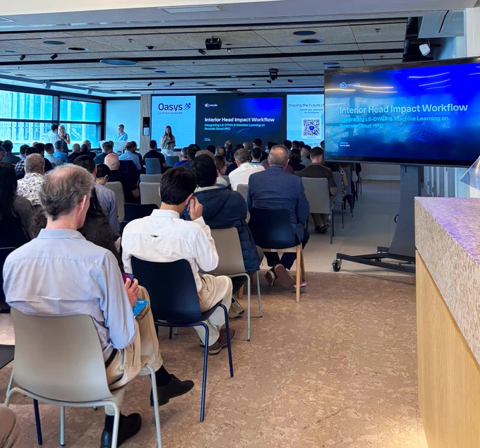 Conference room with attendees seated and facing two large screens displaying a presentation titled “Interior Head Impact Workflow: Integrating LS-DYNA & Machine Learning on Rescale Cloud HPC,” with Rescale and Oasys branding visible. Two presenters stand at the front, and a vertical sign with a QR code reads “Shaping the Future of.”