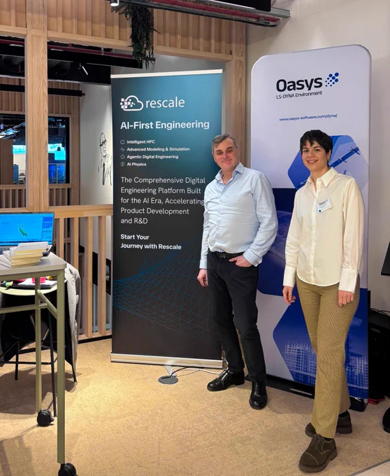 Two people in business-casual attire stand smiling between Rescale and Oasys LS-DYNA Environment banners at an indoor event booth. The Rescale banner reads “AI-First Engineering” with bullets for Intelligent HPC, Advanced Modeling & Simulation, Agentic Digital Engineering, and AI Physics, plus the line “The Comprehensive Digital Engineering Platform Built for the AI Era, Accelerating Product Development and R&D” and “Start Your Journey with Rescale.” A nearby table holds a laptop with a blue screen and papers in a modern, wood-paneled venue. 