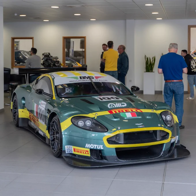 Aston Martin DBR9 race car in green and yellow livery displayed indoors, with Pirelli and Motul branding visible. Attendees stand around the car and in the background, creating a showroom feel during the Le Mans watch gathering.