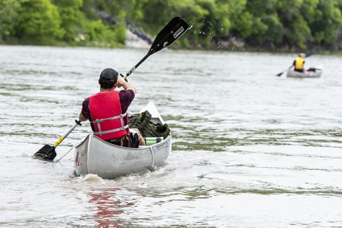 Person wearing a red life jacket paddling a silver canoe on a calm river surrounded by green trees. Personne portant un gilet de sauvetage rouge pagayant un canot argenté sur une rivière calme, entourée d’arbres verts.