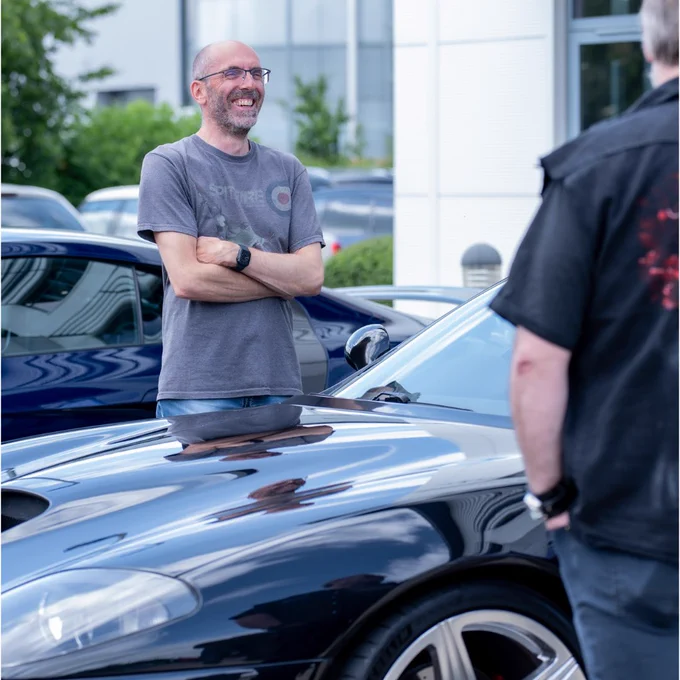 Two attendees talking beside a black Ferrari 550 Maranello, with the front wing and wheel in focus. Other cars sit behind, capturing the informal, enthusiast-led feel of the gathering.