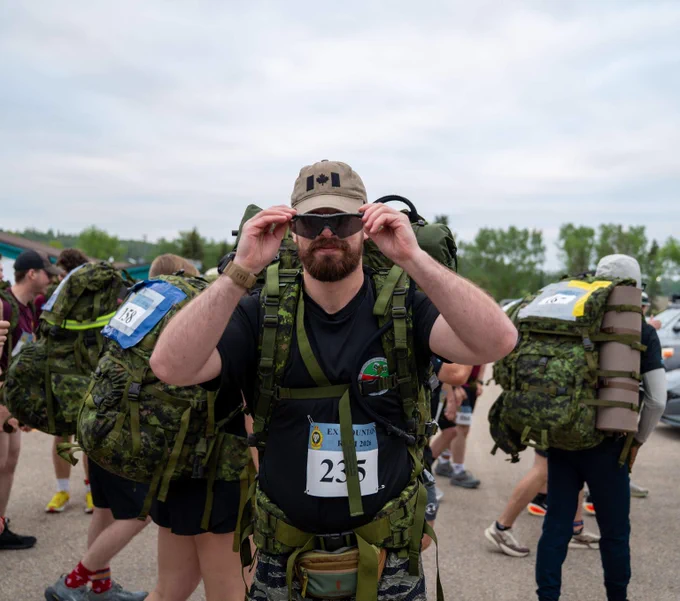 Participant wearing full military gear and a numbered bib adjusting his cap before a group endurance event outdoors. Participant portant un équipement militaire complet et un dossard numéroté ajustant sa casquette avant une épreuve d’endurance en plein air.