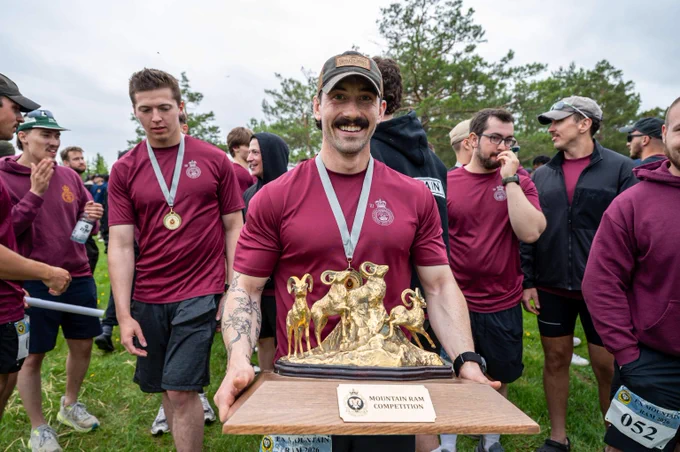 Group of young men in matching maroon shirts celebrating outdoors, one holding a golden ram trophy labeled 'Mountain Ram Competition'. Groupe de jeunes hommes portant des chandails bordeaux assortis célébrant à l’extérieur, l’un tenant un trophée de bélier doré portant l’inscription « Mountain Ram Competition ».