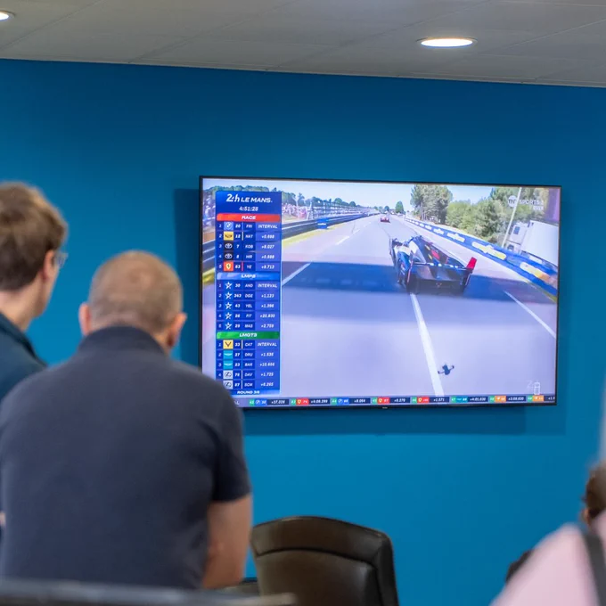 Attendees watching a live broadcast of the 24 Hours of Le Mans on a large screen, with race timing graphics and cars visible on track. The room is set up for a casual watch party with seating and people standing nearby.
