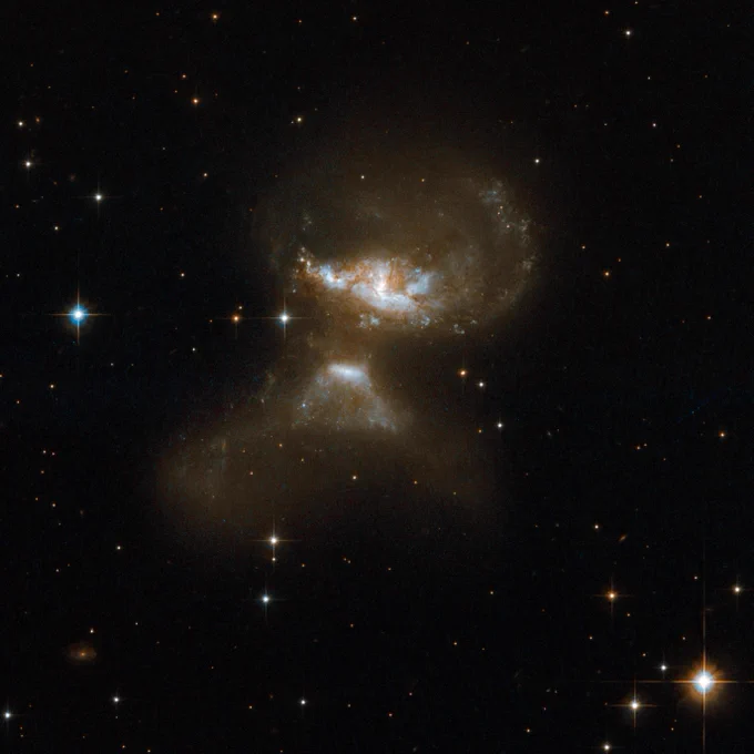 A pair of interacting galaxies shine together against star-studded black space. The two galaxies almost resemble an erupting volcano, with the upper galaxy appearing larger.