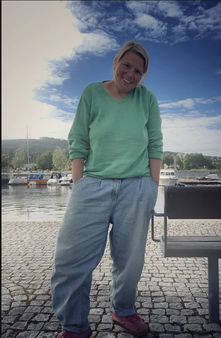 Ich trage eine Barreljeans, sehr weites Bein, am Knöchel umgeschlagen, mit Buntfalten am Bu d, sehr helles Jeansblau und einen grünen MänberFreinstrickpulli mit V Ausschnitt, die Ärmel hochgekrempelt und pinke Vans. Ich stehe auf Kopfsteinpflaster und hinter mir der Hafen mitMotorbooten.