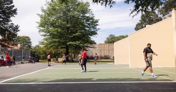 Several people playing handball at the courts in Forest Park as a full-leaf green trees tower over the courts.
