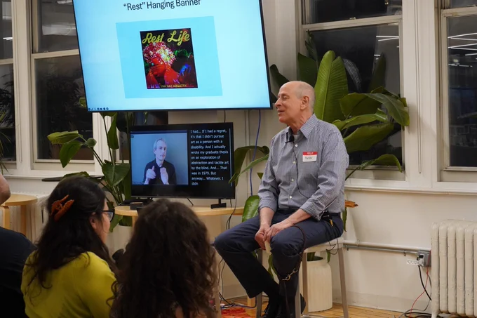 Peter Slatin sits on a stool next to a large screen with his slides and below the screen is a smaller screen displaying an ASL interpreter and captions.