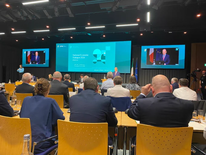 An Taoiseach Micheál Martin addresses delegates at the National Economic Dialogue 2026 (#NED26), hosted by the Department of Finance and the Department of Public Expenditure, Infrastructure, Public Service Reform and Digitalisation. Delegates are seated at round tables in a conference room, with the speaker visible on stage and on large screens to either side.