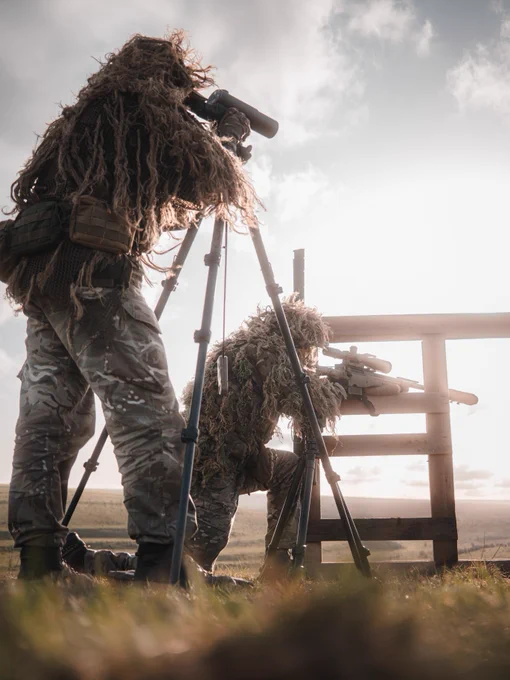 Two individuals in camouflage gear are using binoculars and a rifle with a scope, both positioned on tripods. They are aiming towards a distant landscape under a cloudy sky.