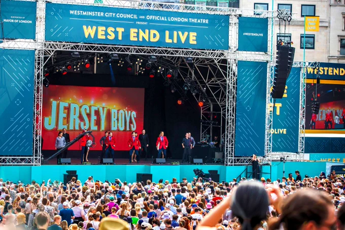 The Jersey Boys on stage at West End LIVE performing against their logo (Jersey Boys written in white on a red background). You can see the heads of hundreds of crowd members. 