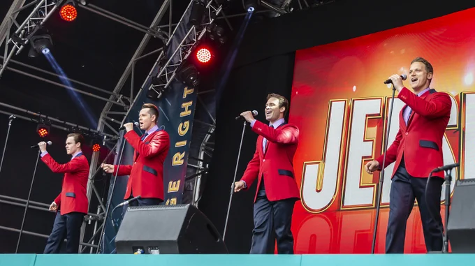A music performance on stage with four people in red jackets singing into microphones. A large screen in the background displays the word "JERSEY." Stage lighting includes red spotlights.