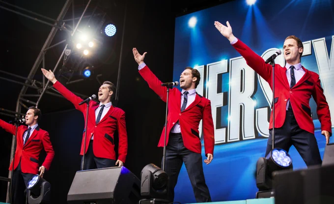 Four performers in red suits sing on stage in front of a "Jersey Boys" sign. They are holding microphones and have one arm raised. Stage lights illuminate the scene.