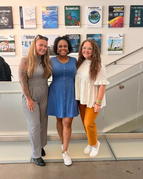 Three women standing together indoors in front of a wall displaying scientific journal covers, dressed in casual outfits with one in a blue dress, another in a gray jumpsuit, and the third in a white blouse and mustard pants.