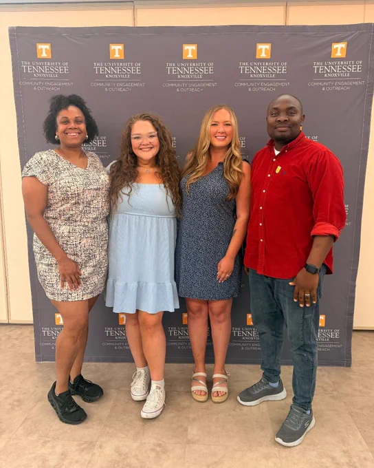 Four students stand side by side in front of a University of Tennessee Knoxville backdrop, dressed in casual summer attire.