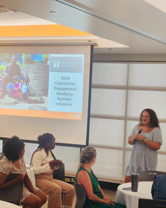 A woman in a gray dress speaks into a microphone during a presentation for the 2026 Community Engagement Academy Summer Intensive while three seated attendees listen attentively.