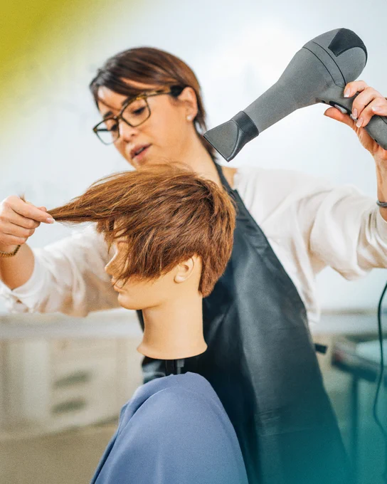 Alt text: A hairstyling student uses a blow dryer and mannequin head to practice hair styling techniques in a salon training environment.