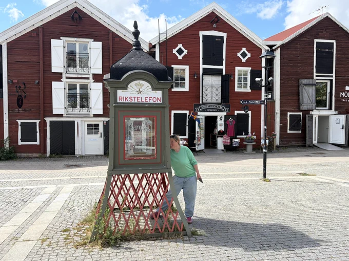 Diese schwedische „Rikstelefon“-Zelle ist der absolute Design-Hauptgewinn! Unten ein weinrotes Holzgitter, das aussieht, als hätte es früher mal als stylischer Gartenzaun gedient, oben ein olivgrünes Häuschen mit schmuckem Goldkrönchen. Das schwarze Prunkdach obendrauf verleiht dem Ganzen den majestätischen Vibe einer Mini-Kathedrale für konsequent analoge Kommunikation. Wer braucht schon 5G, wenn man hier stilecht den Hörer schwingen kann?
Hinter diesem Schmuckstück lugt eine Frau im hellgrünen Pulli hervor, die sich die beste Deckung des Platzes ausgesucht hat. Blöd nur, dass das Smartphone in ihrer Hand sie als modern entlarvt – wahrscheinlich googelt sie gerade insgeheim, wie so ein historischer Kasten überhaupt funktioniert.
Flankiert wird das Spektakel von einer Kulisse aus typisch schwedischen, falunroten Holzhäusern, die so bilderbuchmäßig aussehen, dass man sofort nach Zimtschnecken Ausschau halten möchte. Nostalgie pur mit einem Augenzwinkern!