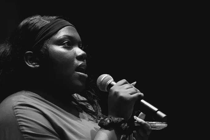 A striking black and white profile photograph of a young woman passionately singing or speaking into a microphone against a dark background, her hand holding the mic stand closely.