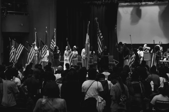 A wide black and white photograph showing a large group of students standing on an auditorium stage holding various American and state flags. An audience of peers sits in the foreground, watching the stage rehearsal.