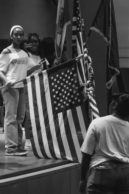 A vertical black and white photograph of two young women standing on a stage holding a large American flag during a rehearsal. One of the women smiles broadly and flashes a peace sign toward the camera while another student looks on from the foreground.
