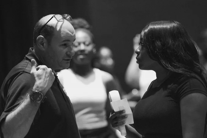 A candid black and white photograph of a man with glasses resting on his head talking and gesturing with his thumb toward a young woman holding papers, capturing a focused moment of event coordination.