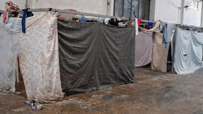 Galpão com estruturas metálicas onde cobertores e panos estão pendurados, formando divisórias. Roupas apoiadas e sandálias no chão.