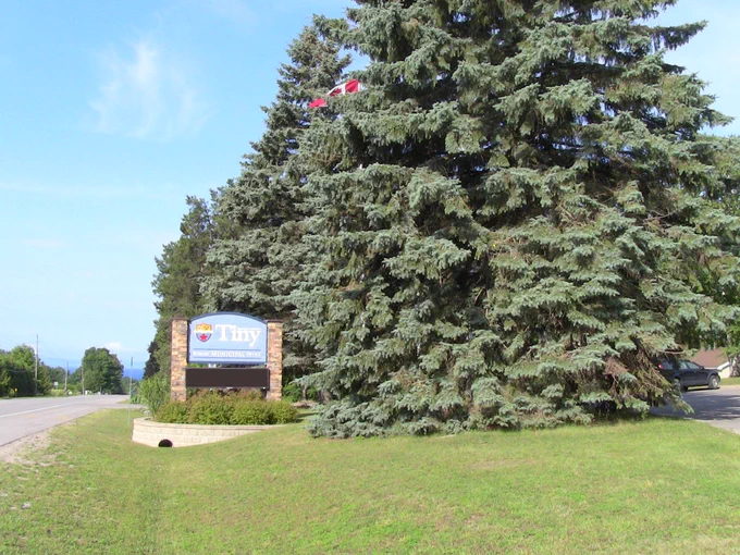 Welcome sign for the Township of Tiny beside a road, surrounded by large evergreen trees under a clear blue sky.
