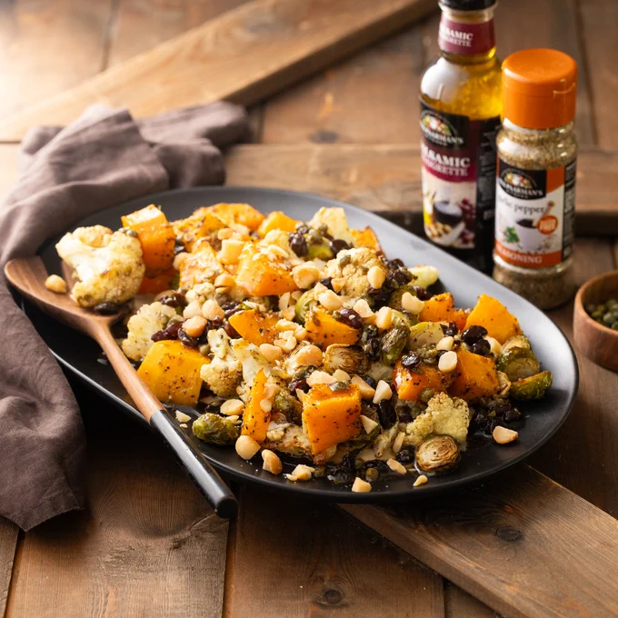 A plate of roasted vegetables, including squash, Brussels sprouts, and cauliflower, topped with nuts. A wooden spoon rests on the plate. In the background, bottles of Colavita balsamic vinegar and Colavita seasoning are visible.