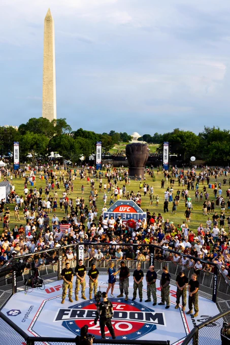 A high-angle view of the UFC Freedom 250 event taking place outdoors on the National Mall in Washington, D.C. In the foreground, a line of military service members stands inside an MMA octagon while a presenter speaks into a microphone. A massive crowd surrounds the cage, with a giant inflatable black fist and the Washington Monument towering in the background.