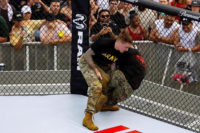 A U.S. Army soldier and a U.S. Marine grappling against the chain-link fence inside a UFC octagon during a mixed martial arts exhibition. The Army soldier, wearing a black \"ARMY\" shirt, has the Marine pressed against the cage while a crowd of spectators watches intently from the other side.