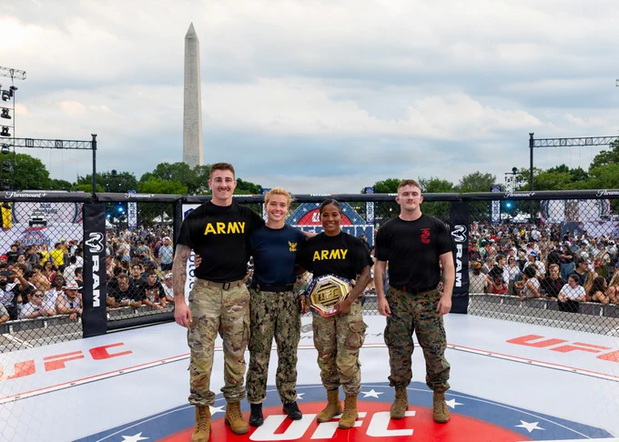 Four U.S. military service members representing the Army, Navy, and Marine Corps smile and pose together inside a UFC octagon. The female Army soldier in the center is proudly holding a UFC championship belt. A large crowd of spectators and the Washington Monument are visible in the background against a cloudy sky.