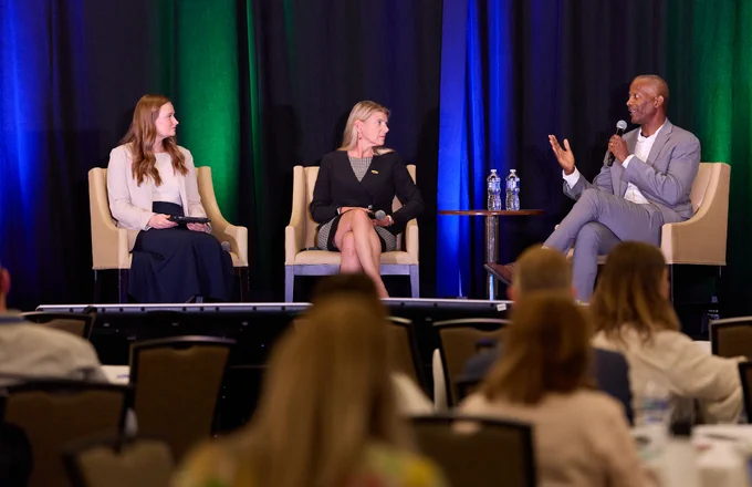 A panel discussion is taking place with three individuals seated on stage, engaged in conversation. One person speaks into a microphone while the others listen attentively. The background features blue and green curtains, and there are audience members visible in the foreground.