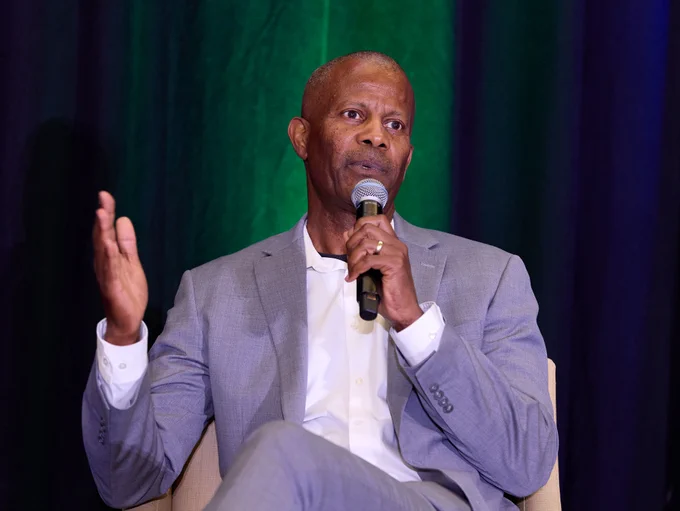A person in a gray suit is speaking into a microphone while seated. There is a dark curtain in the background with green lighting.