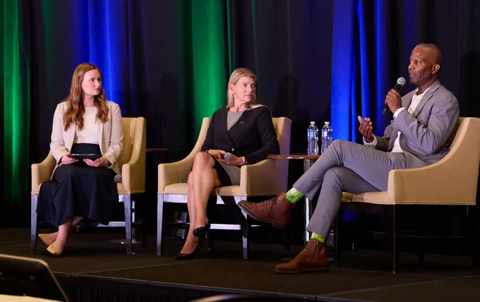 A panel discussion is taking place with three individuals seated on stage. The person on the right is speaking into a microphone. The setting includes a backdrop with green and blue lighting. Bottled water is on one of the tables between them.