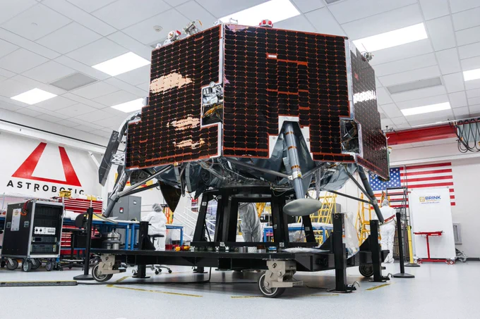 Astrobotic’s Griffin-1 lunar lander sits inside a clean room at the company’s facility. The lander is covered in dark solar panels and mounted on a wheeled test stand. Engineers in white clean-room attire work nearby, with the Astrobotic logo and a large American flag visible in the background. Credit: Astrobotic